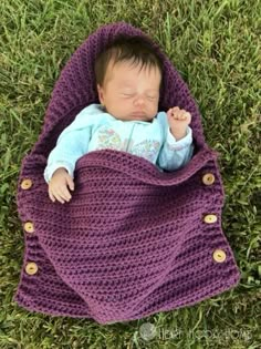 a baby wrapped in a purple blanket laying on top of green grass with its eyes closed