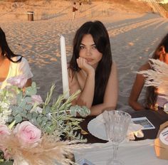 two women sitting at a table with flowers and candles in front of them on the beach