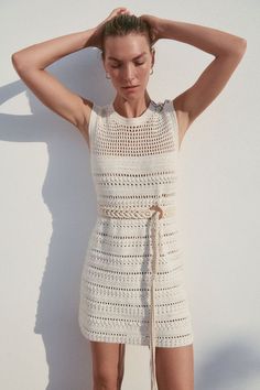 a woman in a white crochet dress with her hands on her head, leaning against a wall