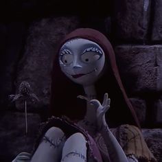 a creepy doll sitting in front of a stone wall with her hand up to the side