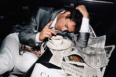 a man sitting at a table with a cup and saucer in his hand while reading the paper