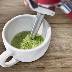 a person using a blender to make green liquid in a white cup on a wooden table