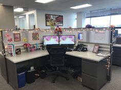 an office cubicle with two computer screens on the desk and lights strung over it