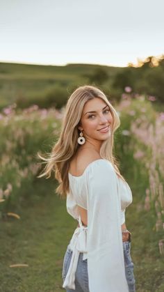 a beautiful blonde woman standing in a field with flowers on the grass and smiling at the camera