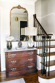 a wooden dresser sitting under a mirror next to a stair case with a lamp on it