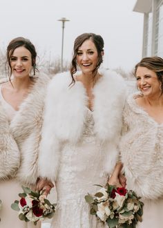 three brides in white fur coats and bouquets smiling for the camera at their wedding