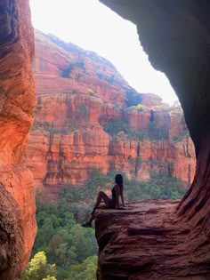 a woman sitting on the edge of a cliff looking out at some trees and mountains