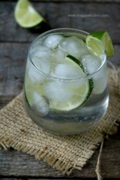 a glass filled with ice and limes on top of a wooden table next to a burlock