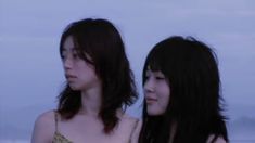 two women standing next to each other on the beach at dusk, one is looking off into the distance