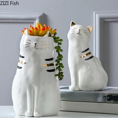 two white ceramic cats sitting next to each other on top of a book shelf with a plant in the middle