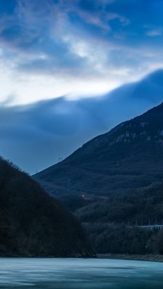 the mountains are covered with trees and clouds in the night time, as seen from across the river