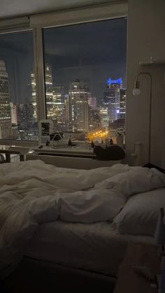 an unmade bed in front of a large window overlooking the city lights at night