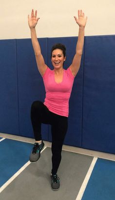 a woman in pink shirt and black leggings standing on one leg with her hands up