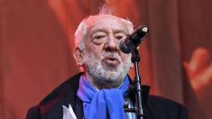 an older man with white hair wearing a blue scarf and holding a microphone in front of him