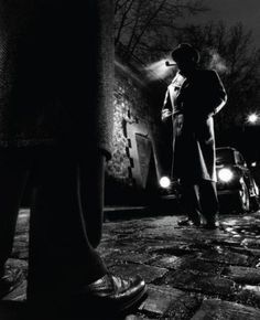 a man standing on the sidewalk at night with his hands in his pockets, looking down