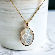 Oval shaped gold Virgin Mary pendant necklace featuring mother of pearl.  Material: Gold plated with cubic zirconia crystals. Oval Shape. Pendant size: 1" x  .5" Chain Length 17" *Gift Box Included 1. Personal Expression of Faith: Wearing this necklace serves as a personal and outward declaration of one's faith. It can act as a daily reminder of one's spiritual beliefs, values, and connection to God. For the individual wearing it, this Christian necklace can also serve as a touchpoint for prayer White Oval Gold Plated Necklace, White Oval Gold-plated Necklace, Gold Oval Mother Of Pearl Necklace, Gold Oval Necklace With Mother Of Pearl, Oval Mother Of Pearl Gold Necklace, White Gold-plated Necklaces For Mother's Day, White Gold Plated Necklaces For Mother's Day, Gold Mother Of Pearl Medallion Necklace, Gold Medallion Necklace With Mother Of Pearl