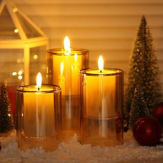 three lit candles sitting on top of snow next to christmas trees and ornaments in front of a window