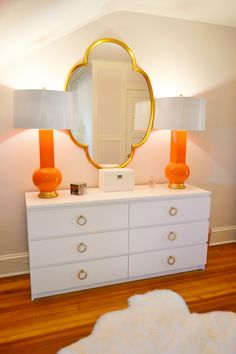 a white dresser with two orange lamps and a gold mirror on it's top