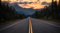 the sun is setting on an empty road in the middle of the woods with mountains in the background