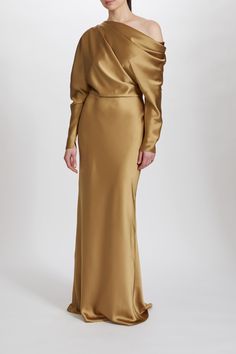 a woman wearing a gold gown with one shoulder and long sleeves, standing in front of a