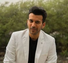 a man in a white suit and black shirt sitting on a bench with trees in the background