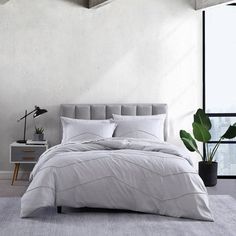 a bed with white sheets and pillows next to a plant in a room that has large windows