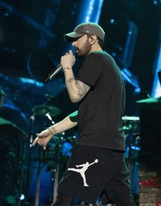 a man in black shirt and cap holding a microphone on stage with other people behind him