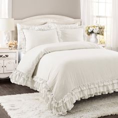 a white bed with ruffles and pillows on top of it in a bedroom