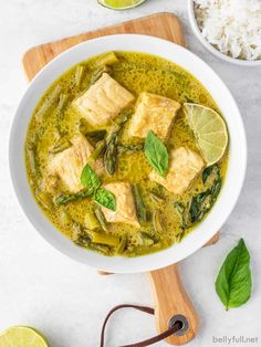 a white bowl filled with fish and green curry next to rice, lime wedges and a wooden cutting board