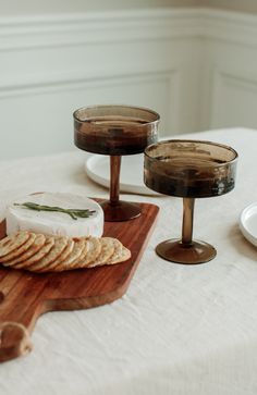 cheese and crackers on a wooden cutting board with wine glasses in the back ground