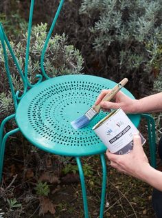 a person painting a chair with paintbrushes in their hands and some plants behind them