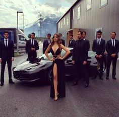 a woman in a black dress standing next to a sports car with men behind her