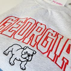 Experience the perfect blend of style and school spirit with our Georgia Bulldog Crewneck Sweatshirt or T-shirt! Available in three colors, this sweatshirt is designed to keep you cozy and comfortable, whether you're cheering in the stands or just showing off your Bulldog pride around town. At the heart of its design is the iconic "Georgia" embroidery, boldly presented across the center chest. The word is intricately stitched in a vibrant red, capturing the essence of the University of Georgia's Georgia Embroidery, Georgia Bulldogs Shirt, Personalized Slippers, Georgia Bulldog, Sewing Seams, Bulldog Shirt, Baby Beanie Hats, Crewneck Design, Baby Bloomers