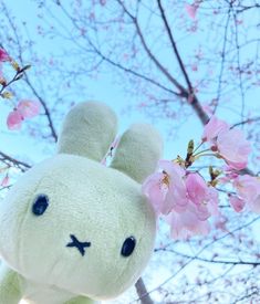 a white stuffed animal with blue eyes standing in front of some pink blossoming trees