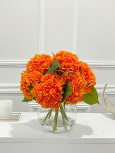 an orange flower arrangement in a clear vase on a white table with candles and other items