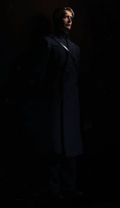 a man standing in the dark with his hands on his hips wearing a black coat