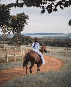 a woman riding on the back of a brown horse down a dirt road next to a field