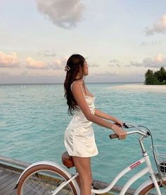 a woman in a white dress standing next to a bike on a pier near the ocean