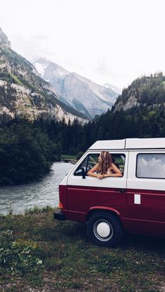 a woman sitting in the passenger seat of a van next to a river and mountains