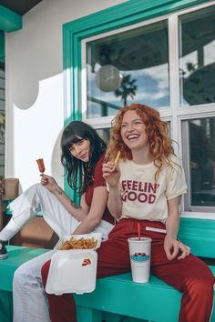 two young women sitting on a bench eating food and drinking soda while one holds a hot dog in her hand