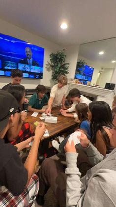 a group of young people sitting around a wooden table in front of televisions on the wall