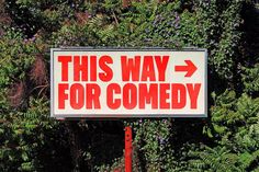 a sign that says this way for comedy in front of some trees and bushes with purple flowers