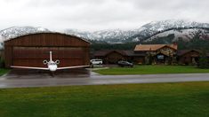 an airplane is parked in front of a large barn with mountains in the back ground