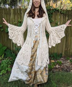 a woman wearing a white crochet hooded jacket and matching skirt is posing in front of a fence