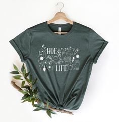 Gardener T Shirt, Plant Lover Shirt, Farmer T Shirt,  Hoeing Ain't Easy Shirt, Gift For Gardeners, Botanical Shirt, Gardening Shirt, Plant Tee Hoe Life Gardening Shirt ❤️  Just in Forrest Heather, but more Colors Soon! Great gifts for anyone. If you like the design, we email an additional discount!  Available from Small to  2 Extra-Large -- ABOUT YOUR NEW TSHIRT -- * 100% combed and ring-spun cotton (Heather colors contain polyester) * Pre-shrunk fabric * Side-seamed construction * Shoulder-to-shoulder taping -- ADDITIONAL NOTES -- This product is made especially for you as soon as you place an order, so we cannot accept returns. Pls, double check your sizing! -- HAVE QUESTIONS or NEED MORE HELP? -- Please message me through Etsy; I'd love to help! Relaxed Fit Shirt With Graphic Print For Gardening, Relaxed Fit Graphic Print Shirt For Gardening, Crew Neck Cotton Shirt For Gardening, Green Tops With Plant Print For Gardening, Green Plant Print Tops For Gardening, Green Tops With Letter Print For Gardening, Green Relaxed Fit T-shirt For Gardening, Botanical Shirt, Gardening Shirts