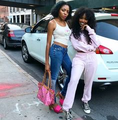 two women standing next to each other on the side of a road with cars behind them