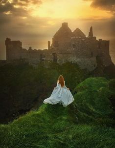 a woman sitting on top of a lush green hillside next to a castle under a cloudy sky