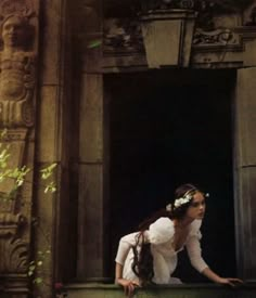 a woman in white dress sitting on the ground next to an open door with flowers in her hair