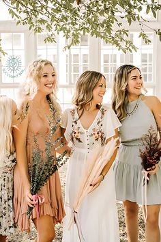 three beautiful women standing next to each other in front of a tree and holding flowers