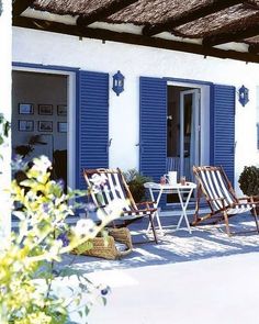 two lawn chairs sitting on the outside of a white building with blue shutters and potted plants